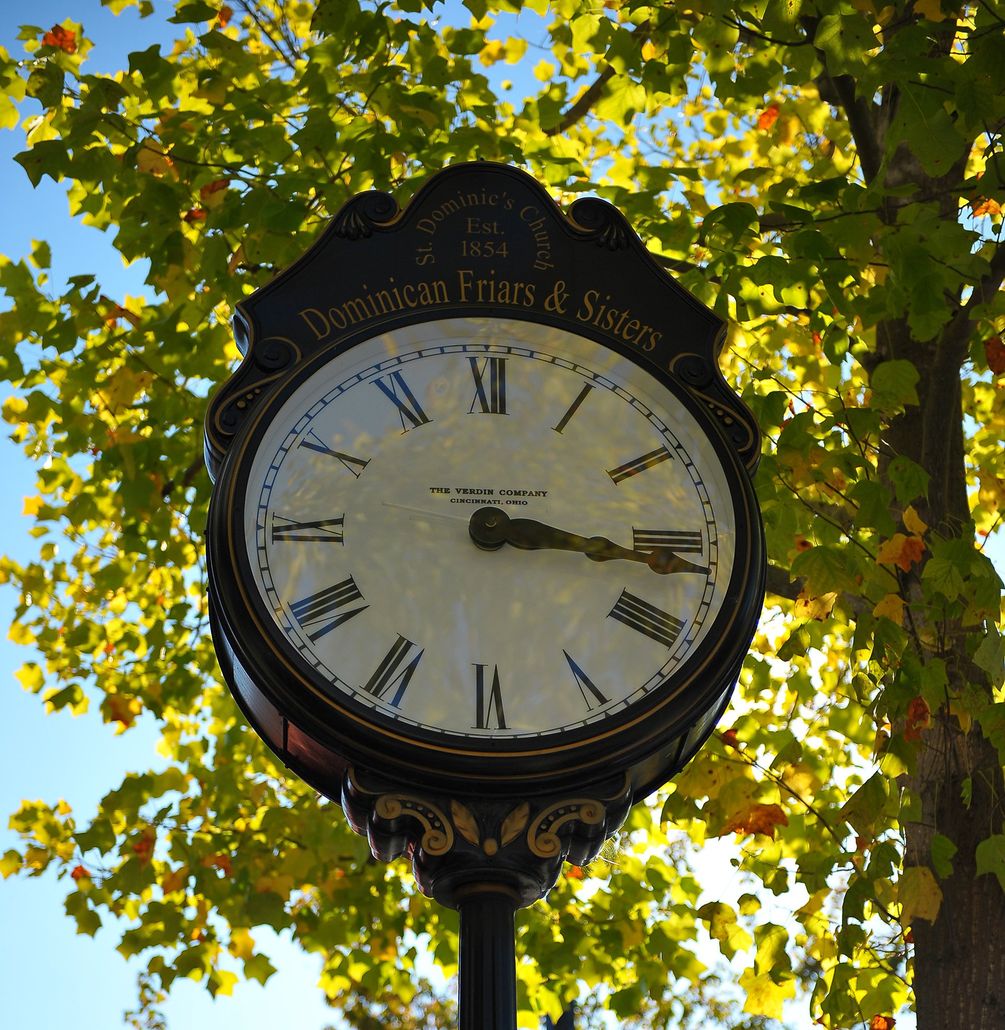 Photo of interesting old clock with roman numerals installed as a prominent sidewalk feature, with green leaves of a tree in the background
