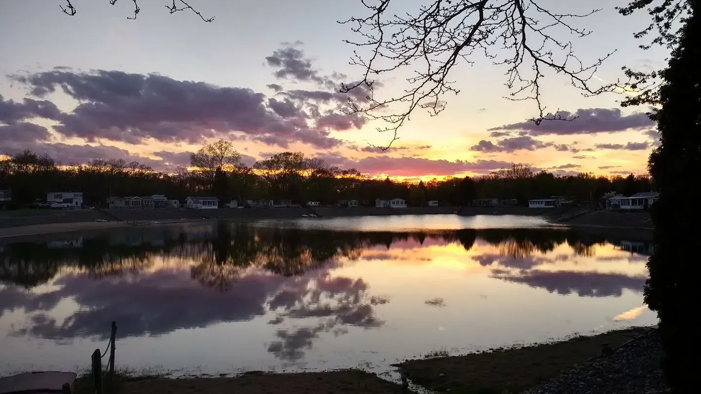Sunset over a calm lake reflecting colorful clouds and trees; small buildings in the distance.