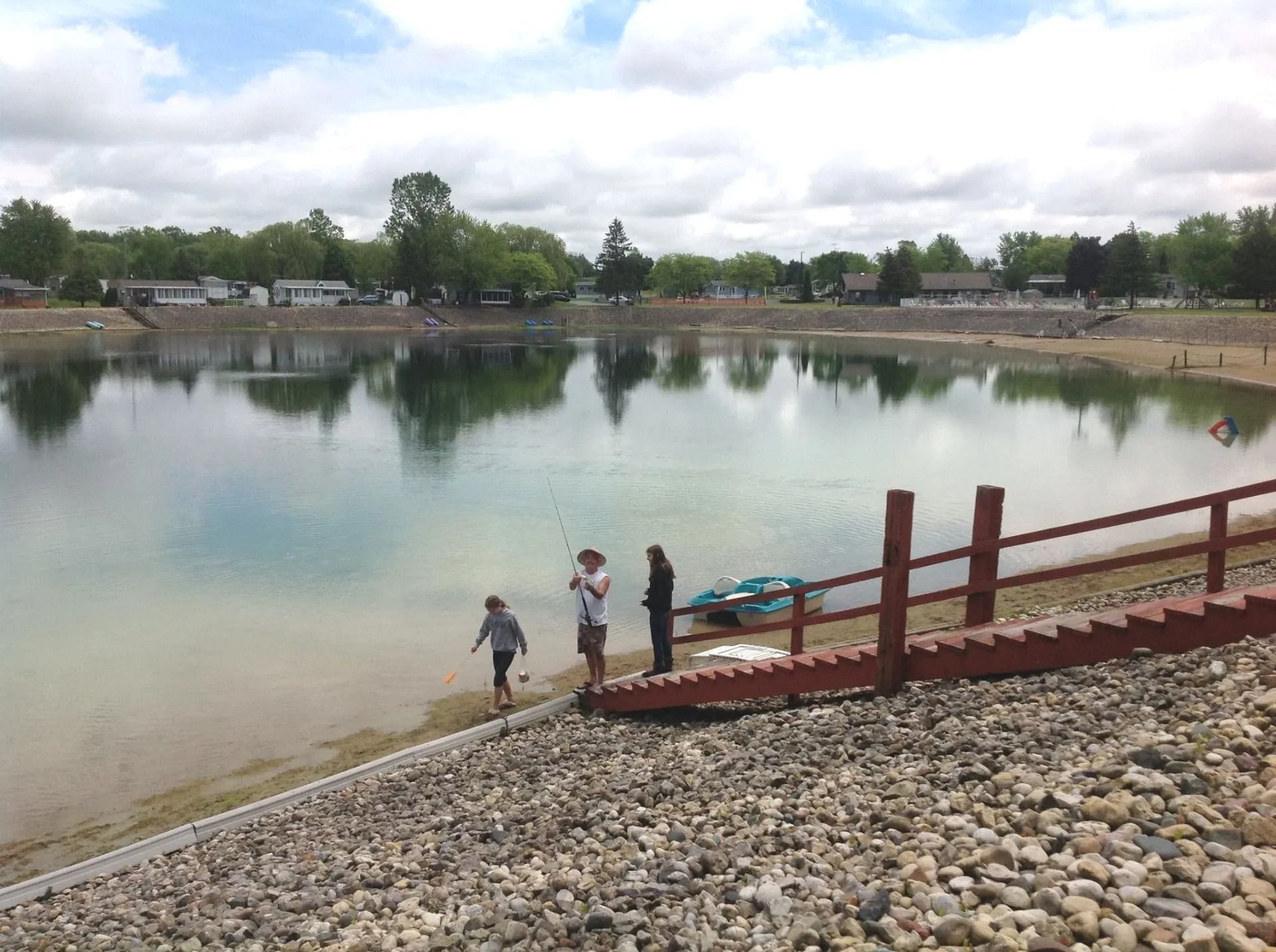 People fishing from a pier at a calm lake on a sunny day. Houses in the background.