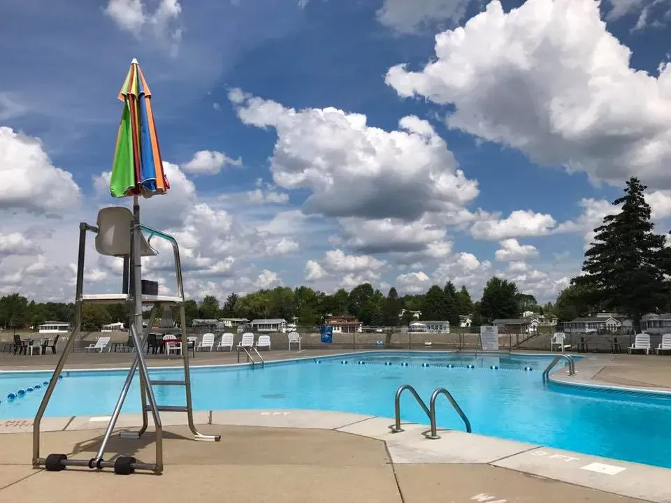A lifeguard chair with a colorful umbrella stands by a blue swimming pool under a bright, cloudy sky.