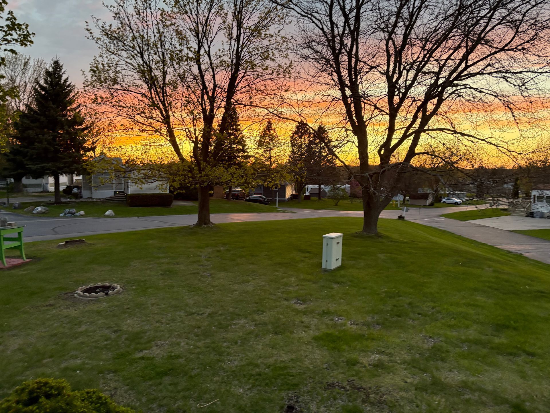 A vibrant sunrise paints the sky in shades of orange and yellow over a grassy suburban neighborhood with trees.