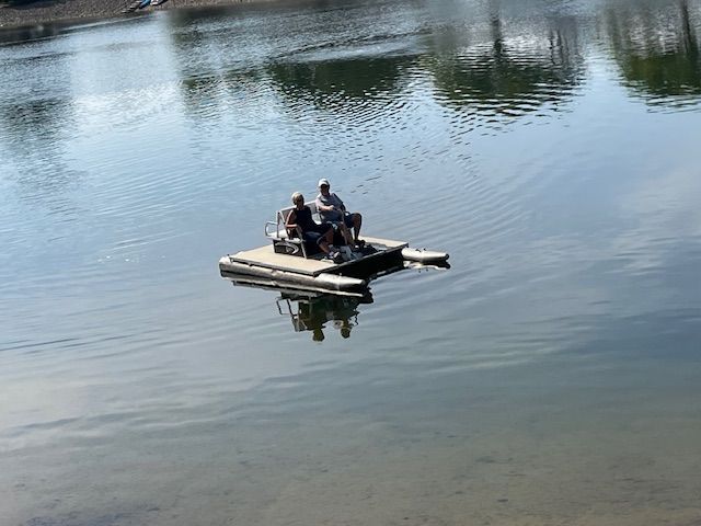 Two people sit on a small, rectangular floating platform on a calm lake, engaged in fishing.