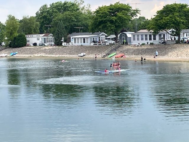 People on a paddleboard in a lake in front of white mobile homes on a sandy shoreline with trees in the background.