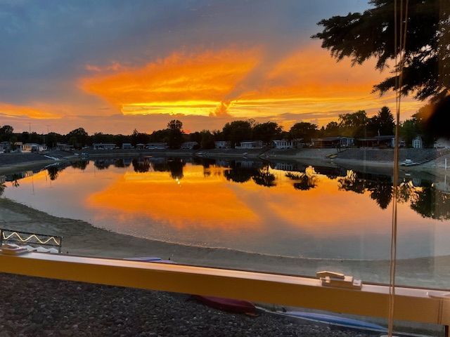 A vibrant orange sunset reflects on a calm lake, bordered by trees and houses, viewed from behind a yellow railing.