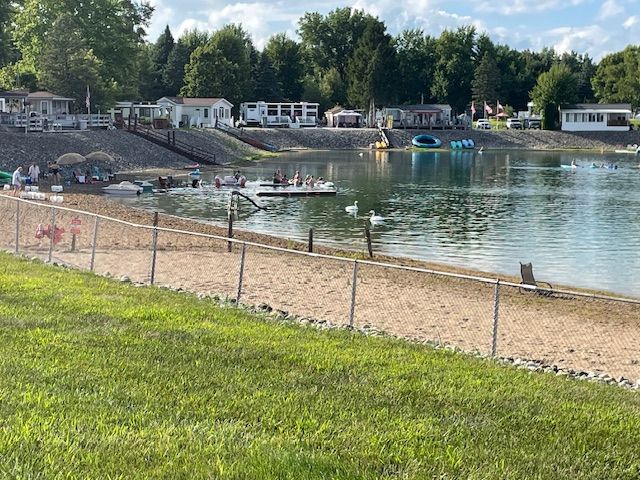 A sandy beach bordering a lake with swimming platforms, swans in the water, and mobile homes on the grassy bank above.