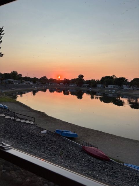 A calm lake reflects a vibrant orange sunset, with small boats resting on the sandy shore in the foreground.