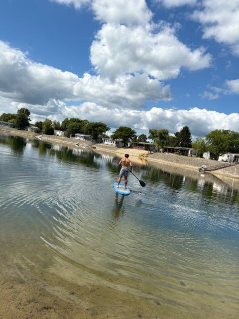 A person stands on a blue paddleboard in a calm lake, using an oar under a bright, cloudy sky with trees and homes nearby.