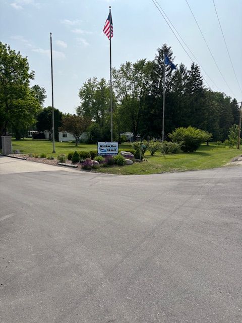 A sign for "Willow Park" stands on a grassy lawn with three flagpoles flying flags under a bright blue sky.