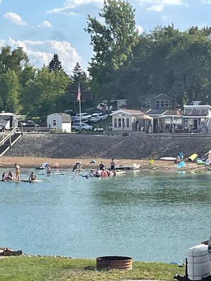 People swim and lounge in a lake near a sandy beach with small waterfront buildings and trees in the background.