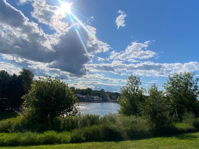 Sunlight shines over a lake, surrounded by green trees and shrubs under a blue, cloudy sky.