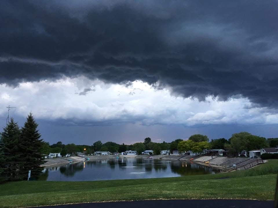 Dark, heavy storm clouds hang over a small pond and a residential area with trees and grassy banks.