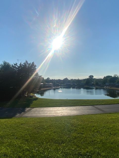 A sunlit lake reflecting the sky, surrounded by green grass and a dark paved path in a quiet, natural park setting.