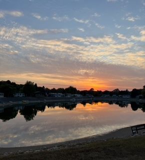 Sunset reflects on a calm lake, with silhouettes of trees and distant buildings under a soft, multi-colored sky.
