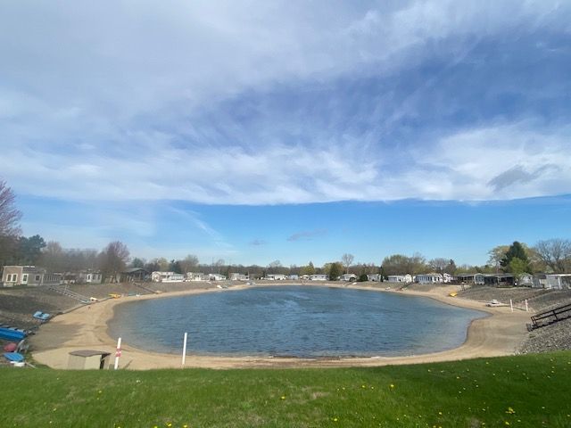 A small, calm pond surrounded by a sandy beach and a ring of mobile homes under a clear, bright blue sky.