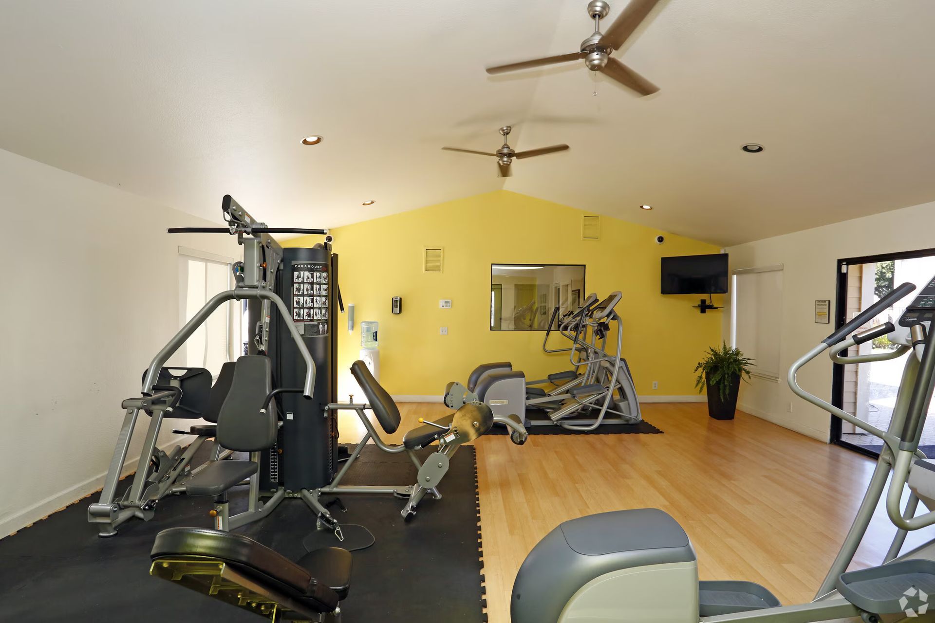 Bright gym room with exercise machines, mirrors, and wooden floors, featuring yellow accent walls.