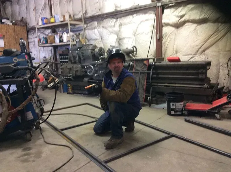 A man is kneeling down in a garage with a welding machine in the background.