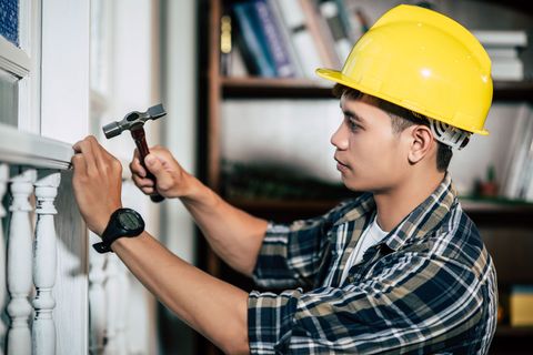 Person wearing a yellow hard hat hammering a nail into white trim.