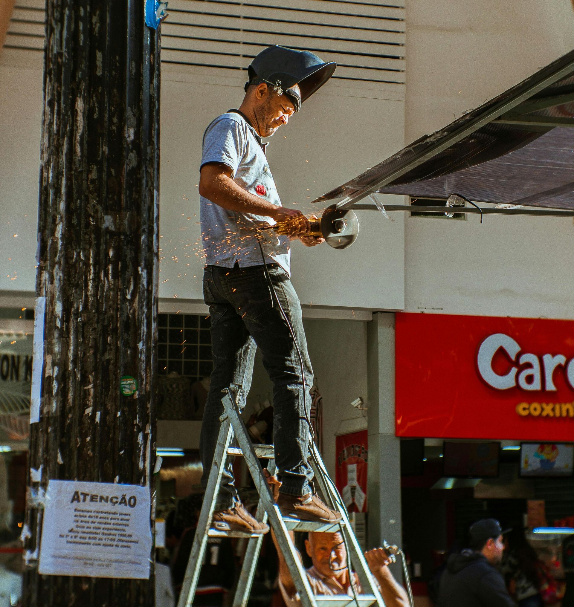 Man on ladder using power tool, sparks flying. Another man steadies ladder near storefront with red sign.