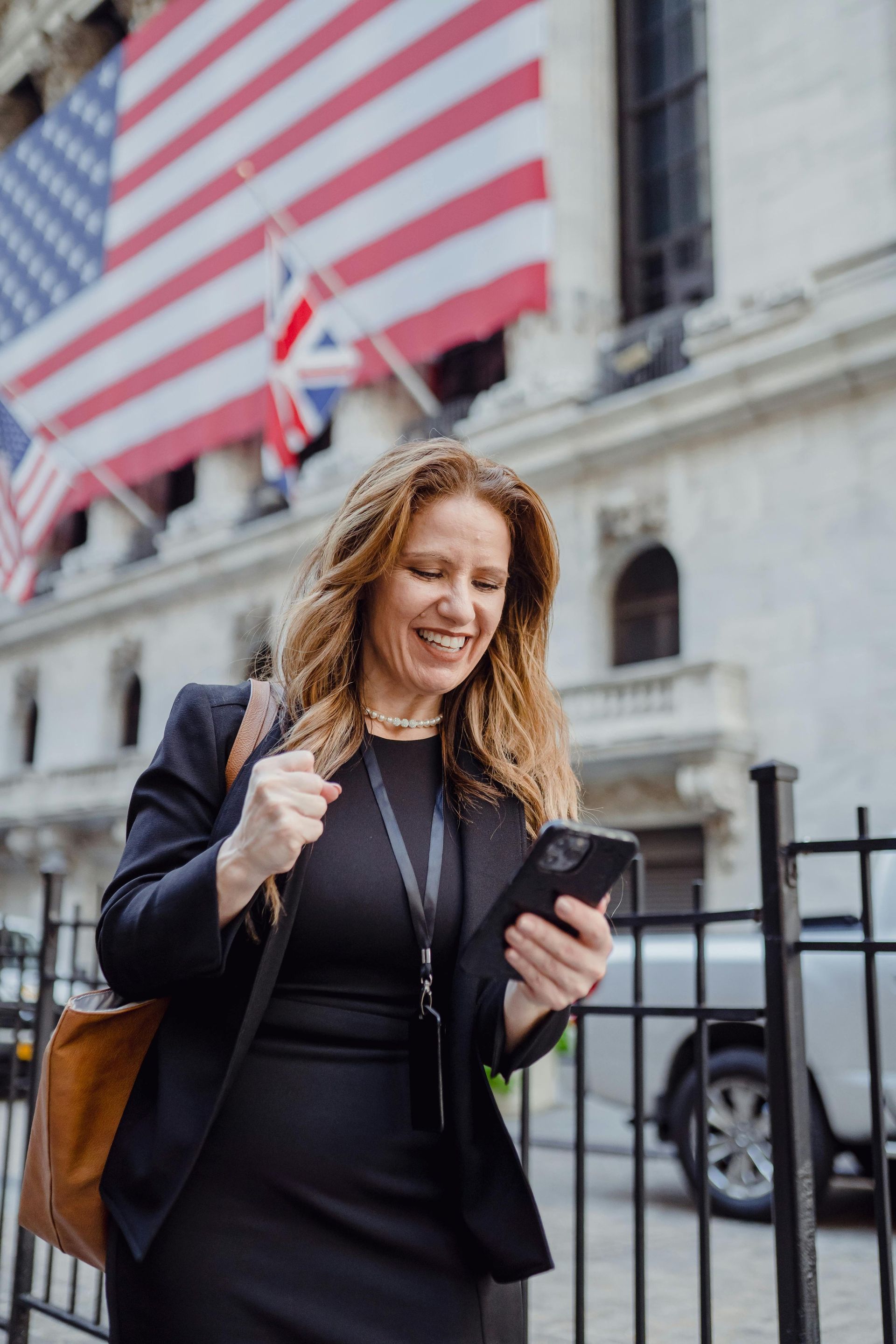 Woman in suit smiles, looks at phone, fists clenched, in front of Wall Street building with US flag.