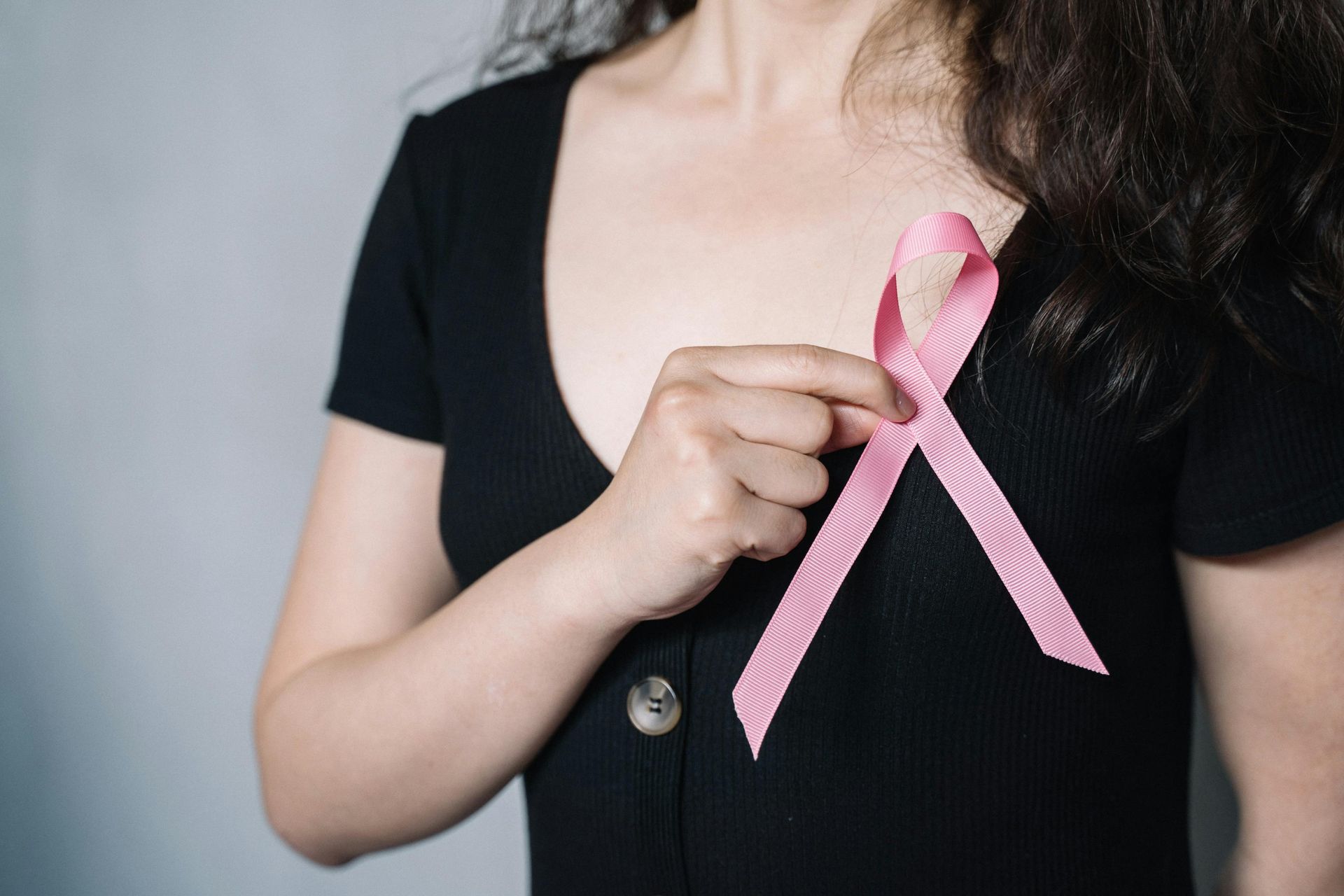 Woman in black shirt holding a pink ribbon, symbolizing breast cancer awareness.