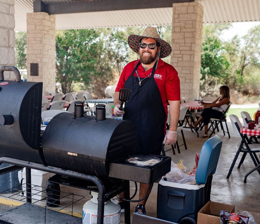 A man wearing a hat and apron is standing in front of a grill volunteering at at the 'Hill Country Cup'