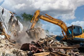 Yellow excavator demolishing a building; debris and dust in the air under a cloudy sky.