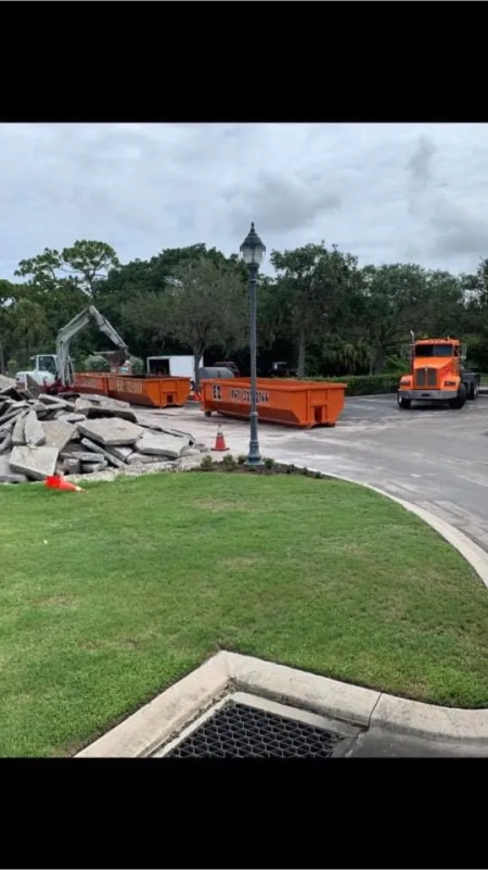 Construction scene: orange dumpsters, truck, and excavator on paved road near a lawn.