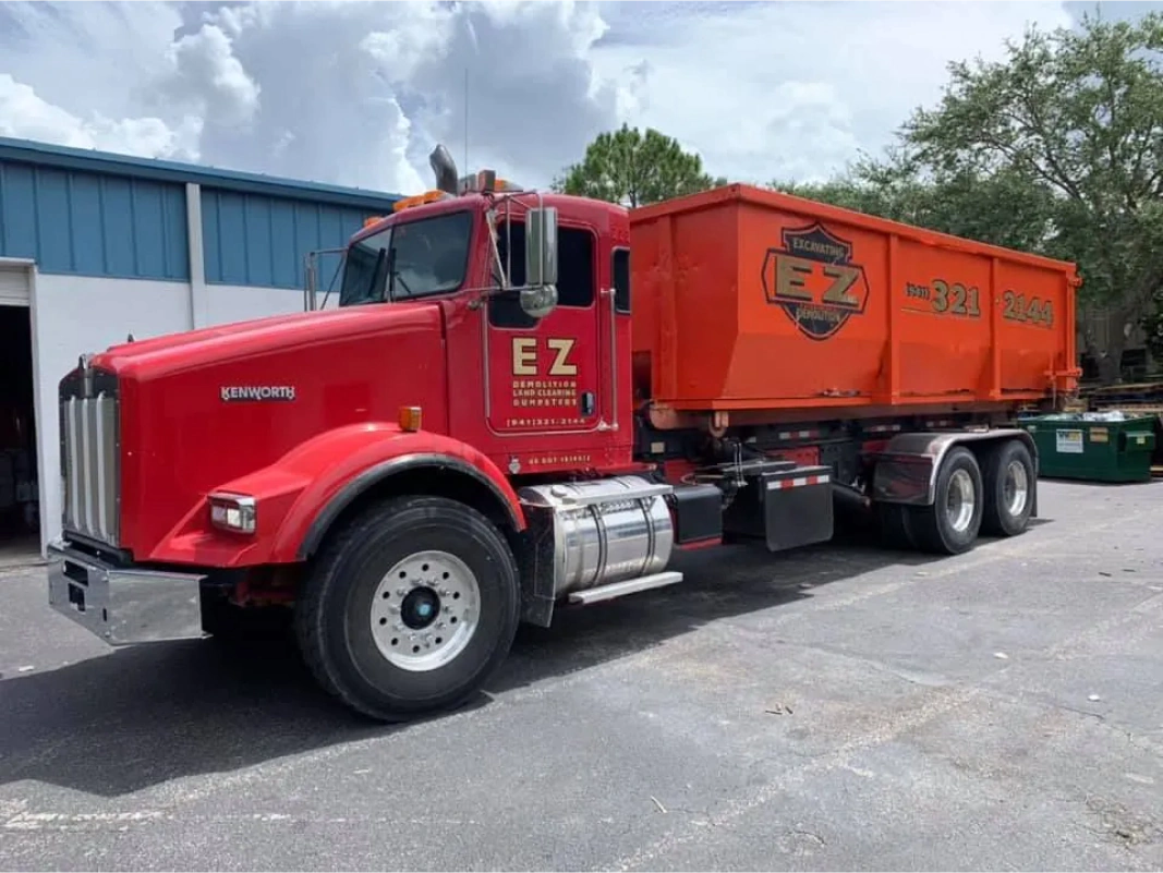 Red Kenworth dump truck with orange EZ Dump dumpster, parked outside a building.