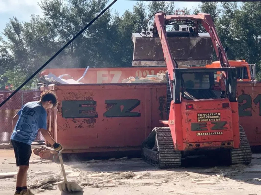 Man shovels debris near orange dumpster, with a skid steer loader dumping material.