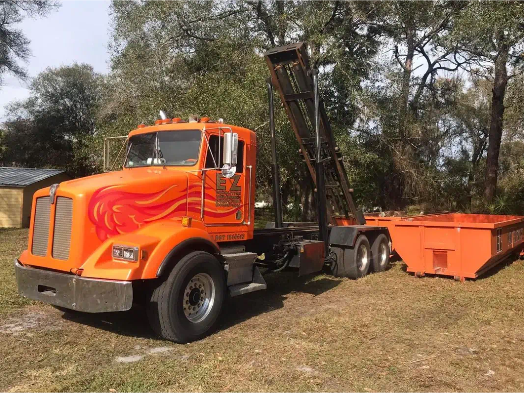 Orange roll-off truck with matching dumpster in a grassy area, trees in background.