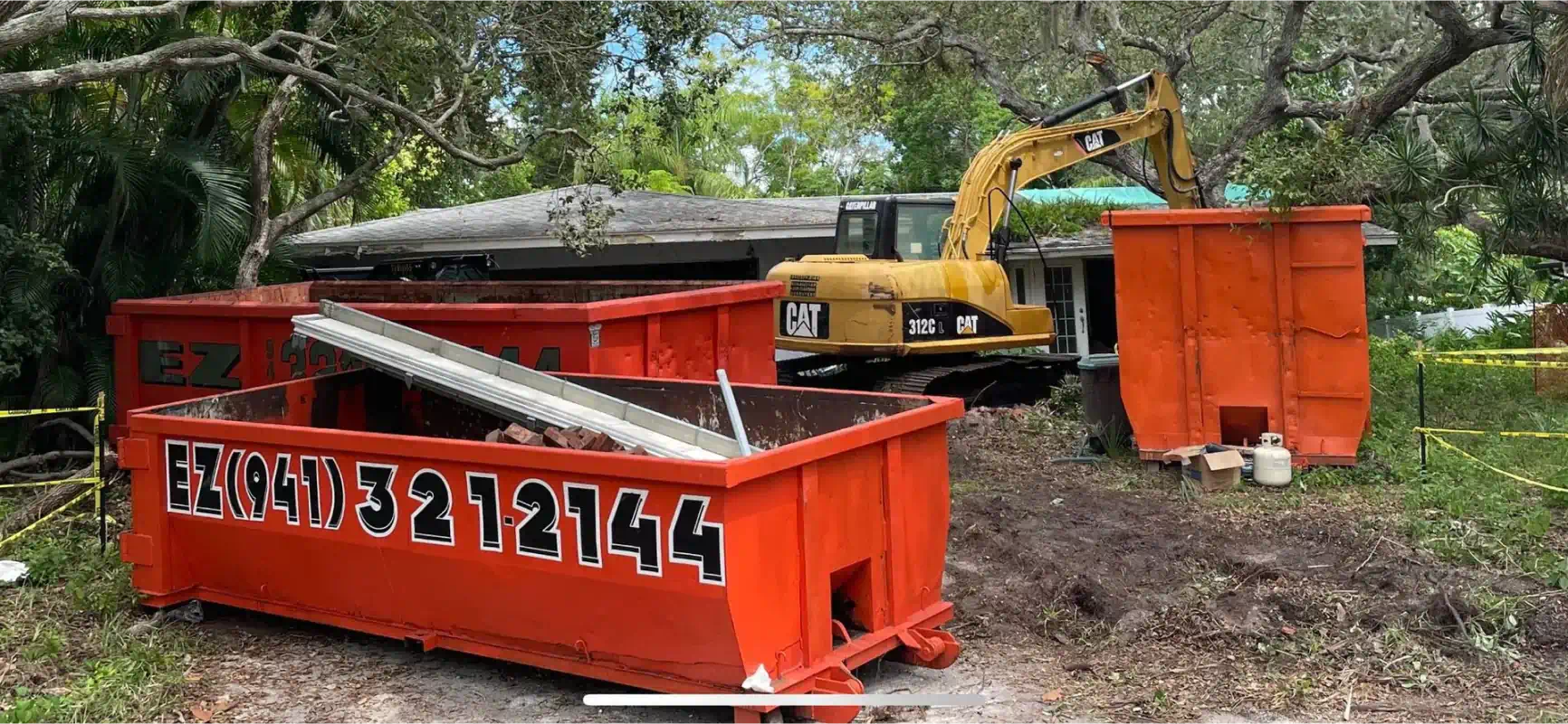 An excavator demolishes a house. Red dumpsters are in front.