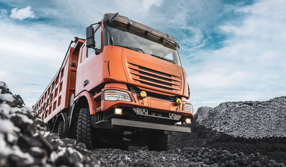 Orange dump truck on a pile of dark rocks under a cloudy blue sky.