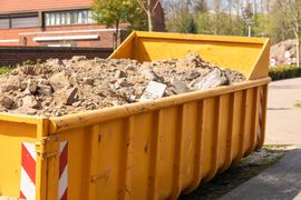 Yellow dumpster filled with dirt and debris, parked on a street near a brick building.