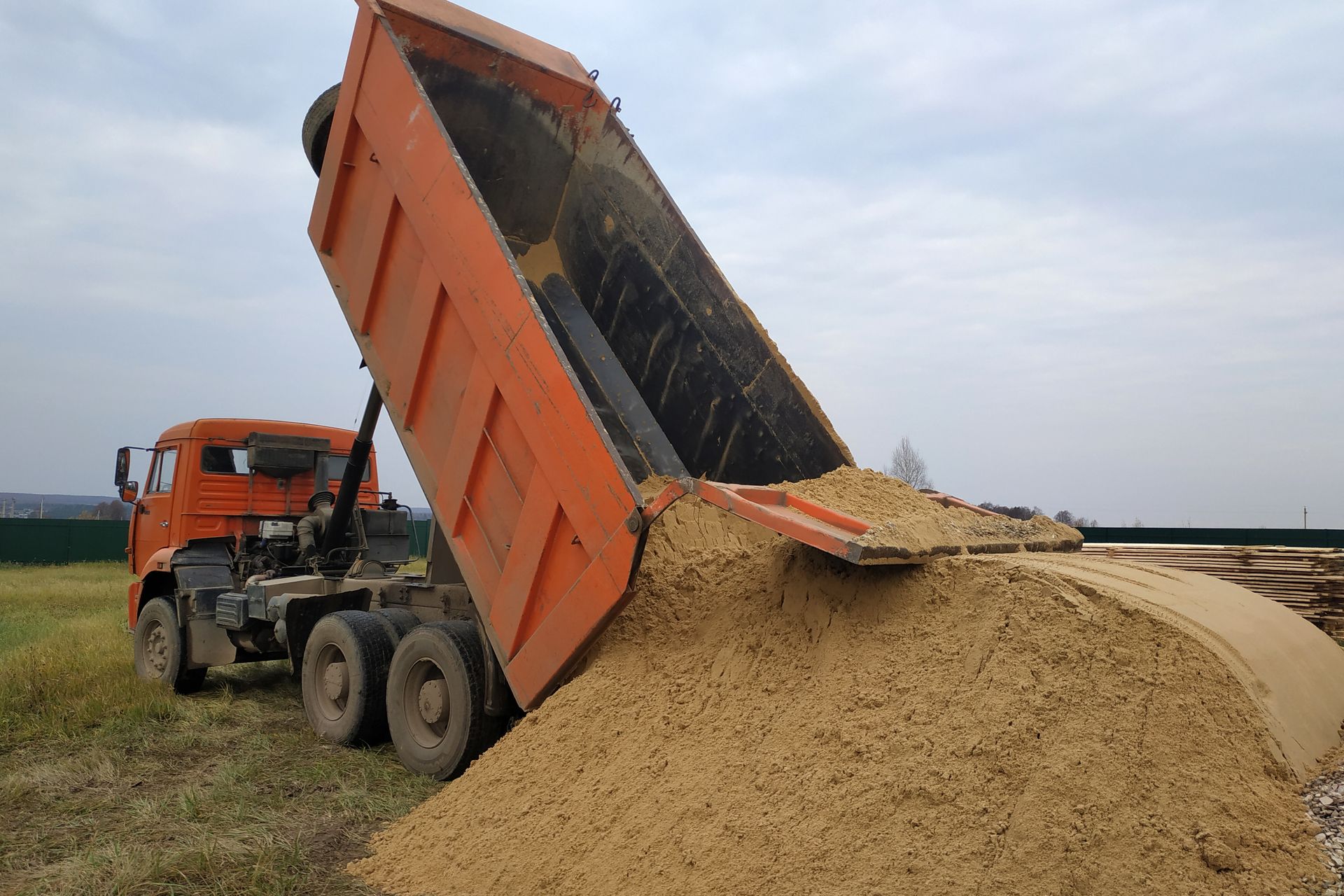 Orange dump truck unloading sand on a grassy field under a cloudy sky.