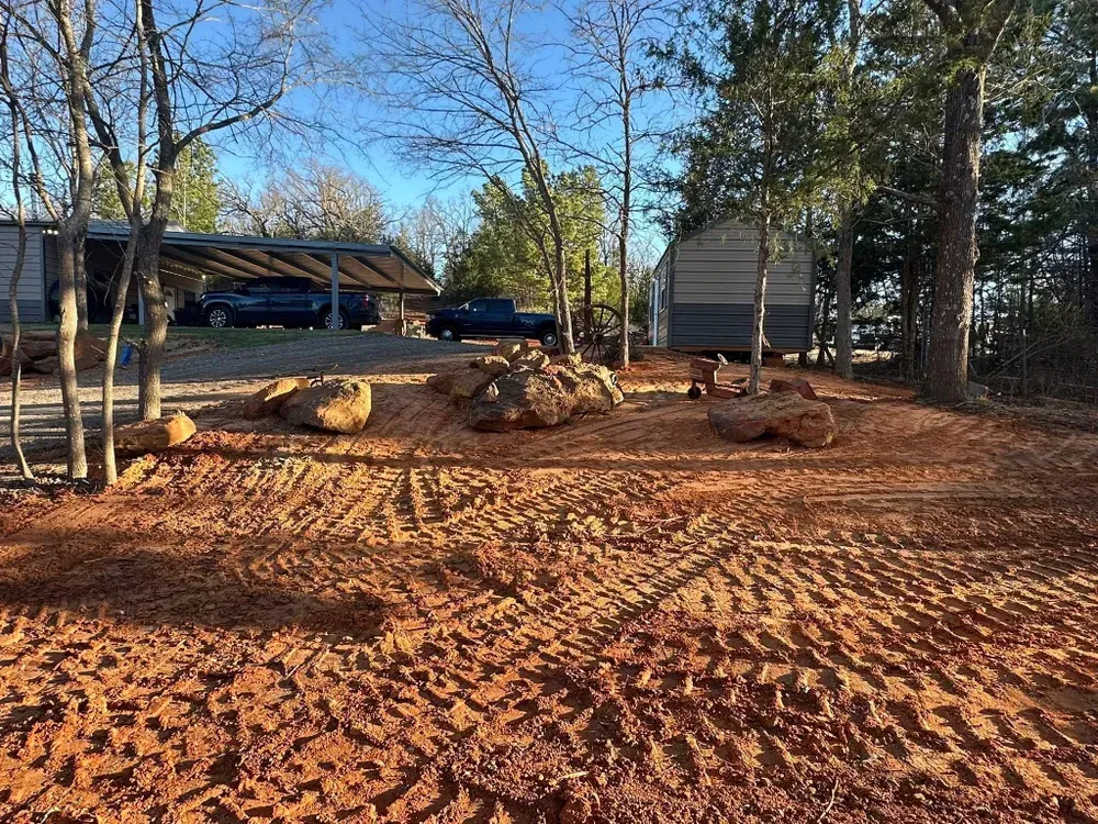 Dirt lot with tire tracks, scattered rocks and trees beside a house under a blue sky.