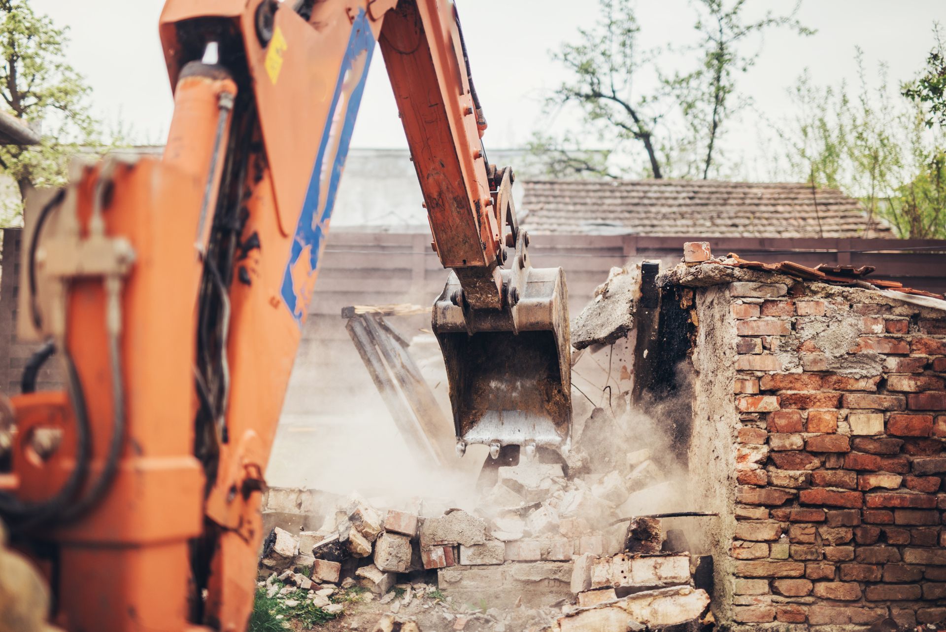 Orange excavator demolishing a brick building, with dust rising from the rubble.