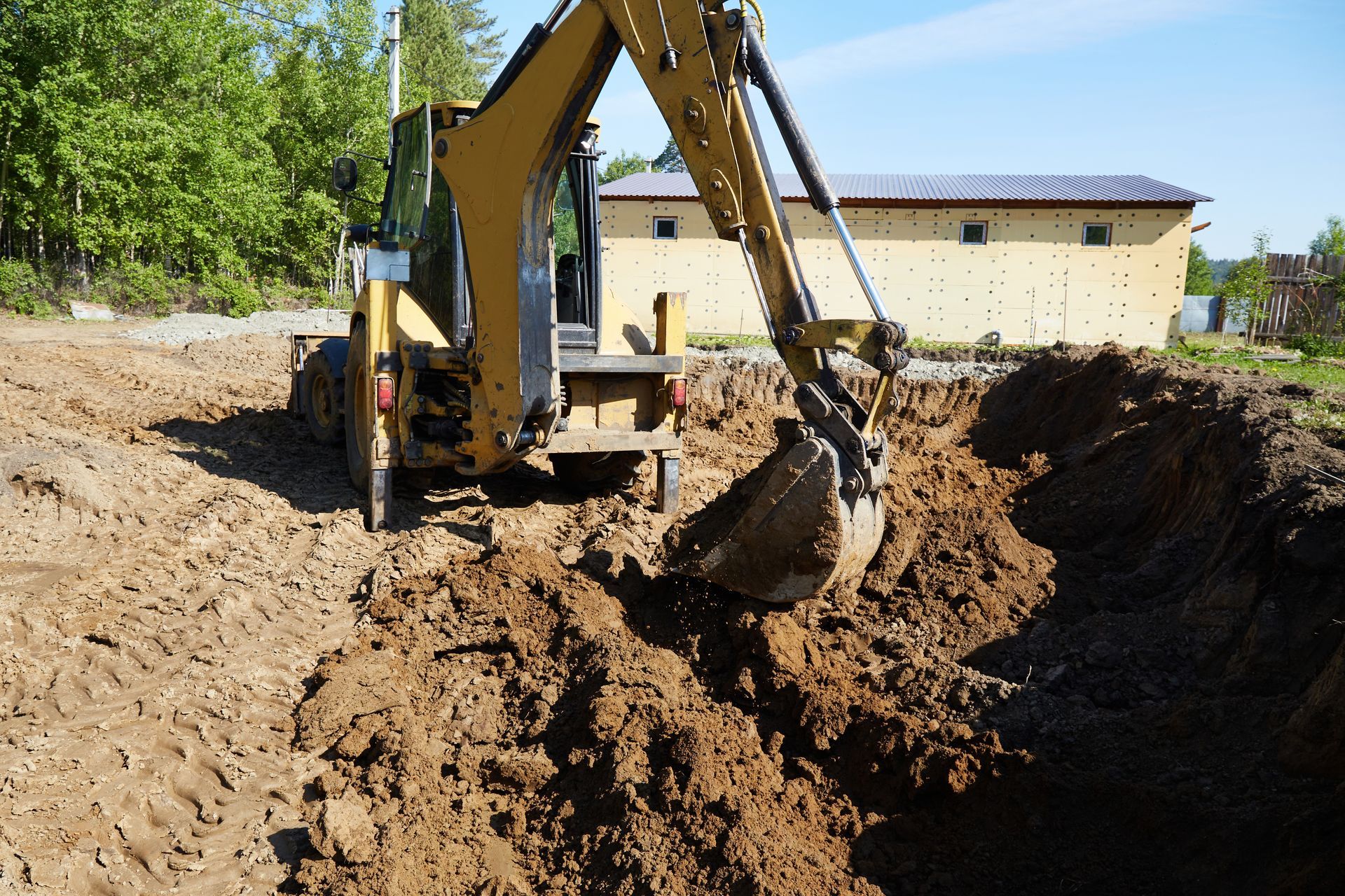 Excavator digging a large dirt trench beside a building on a sunny day