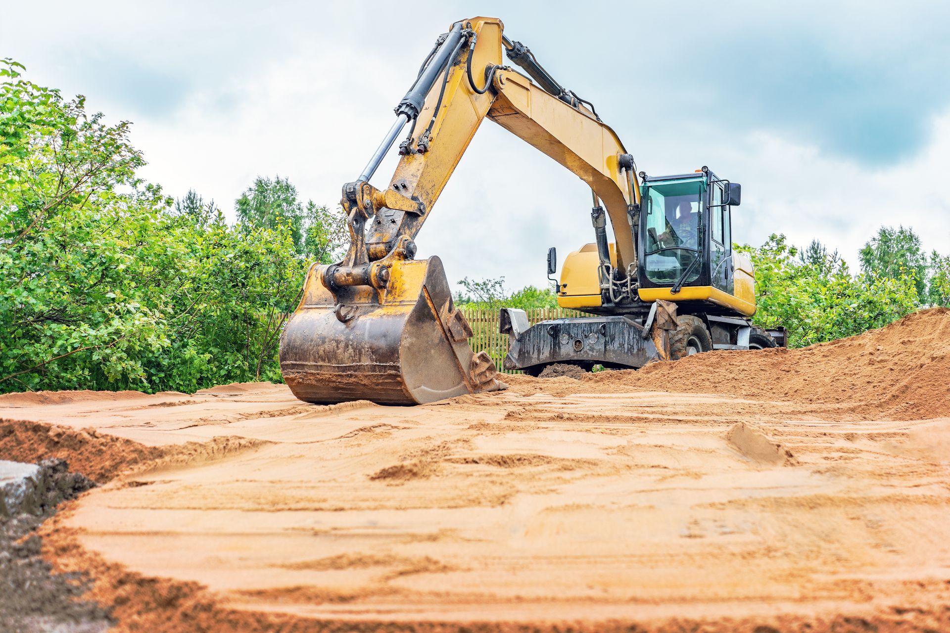 Yellow excavator grading a sandy dirt road beside green trees under a bright sky