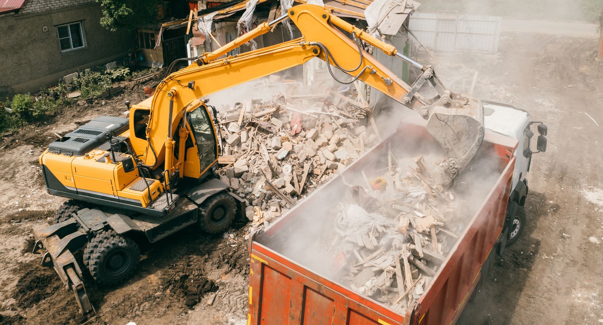 Excavator loading rubble into an orange dumpster at a demolition site, with dust rising.