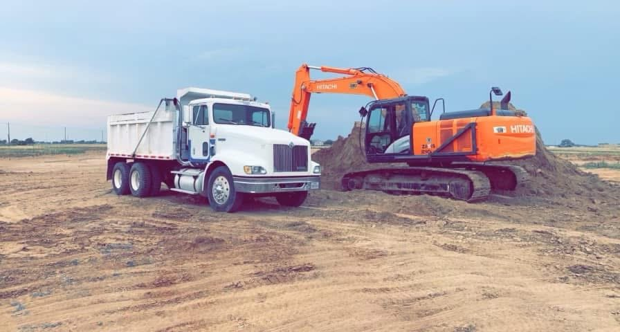 Turquoise excavator digging dirt at a construction site with cranes and buildings in the background