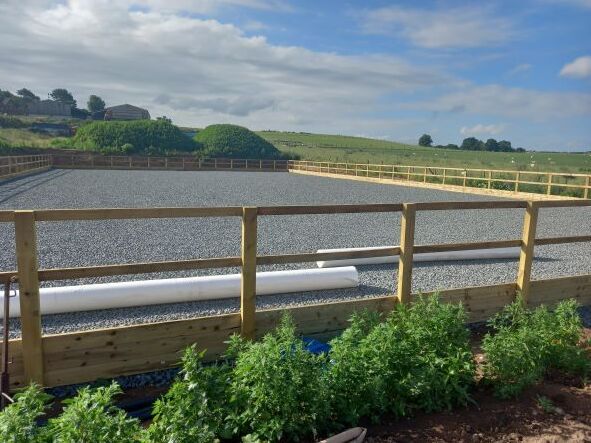 A wooden fence surrounds a gravel area in a field.