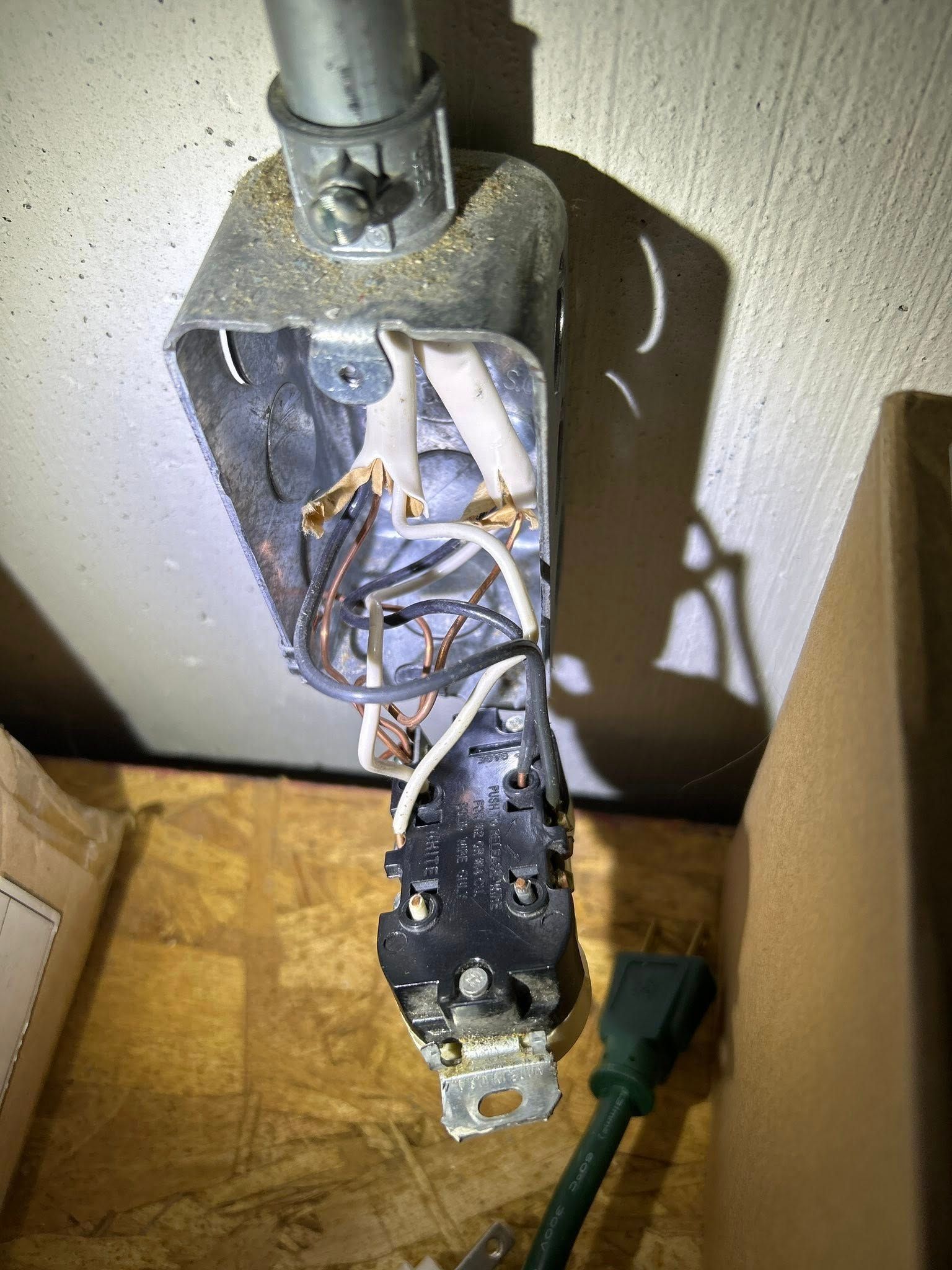 Open electrical outlet box with exposed wires, a connected green power cord, and a metal conduit.