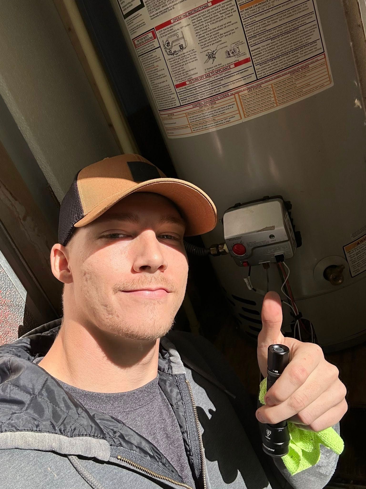 Man giving thumbs-up next to water heater, holding a flashlight. Brown hat, work clothes.