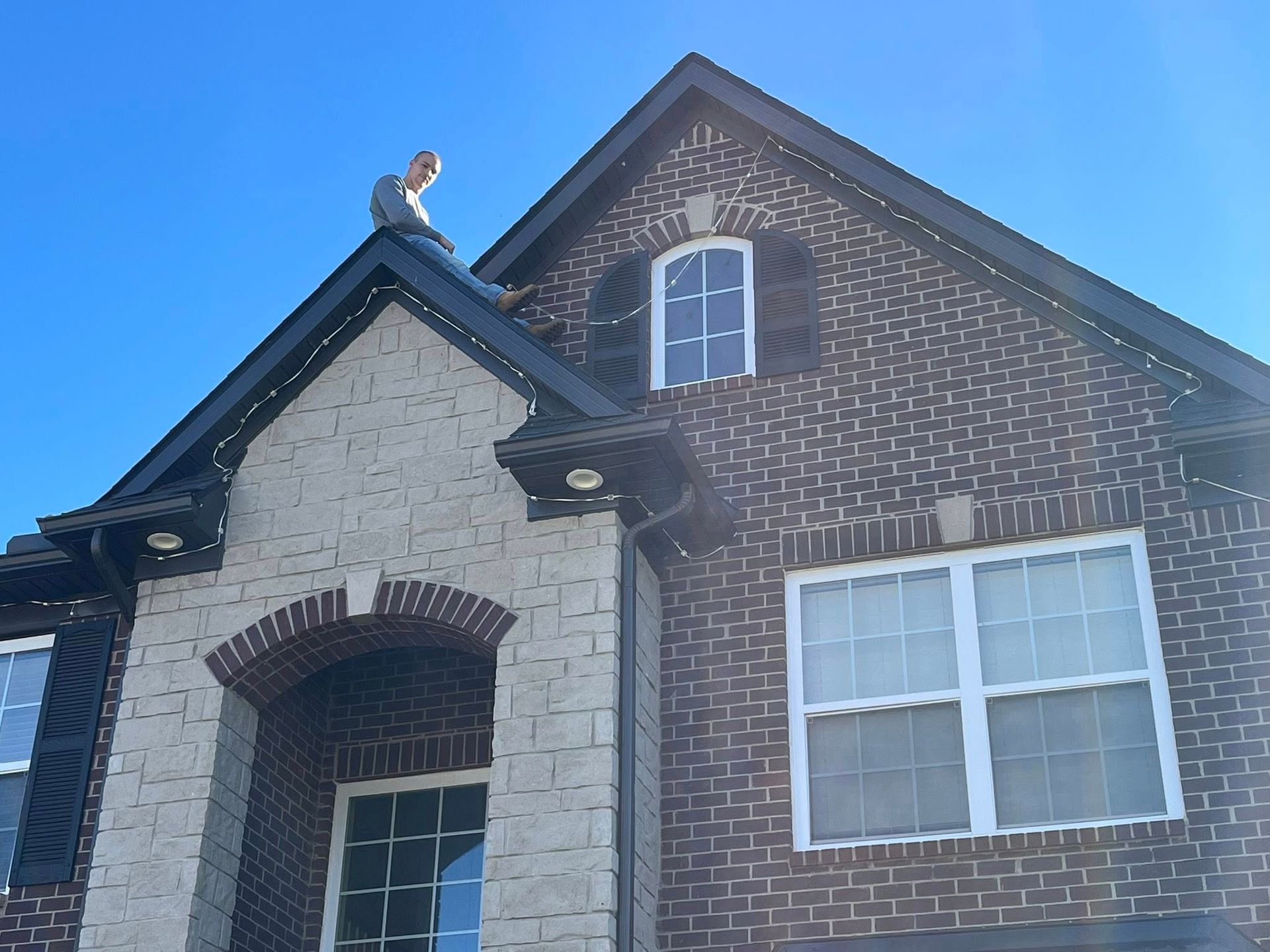 Person on a roof installing lights on a brick house against a blue sky.