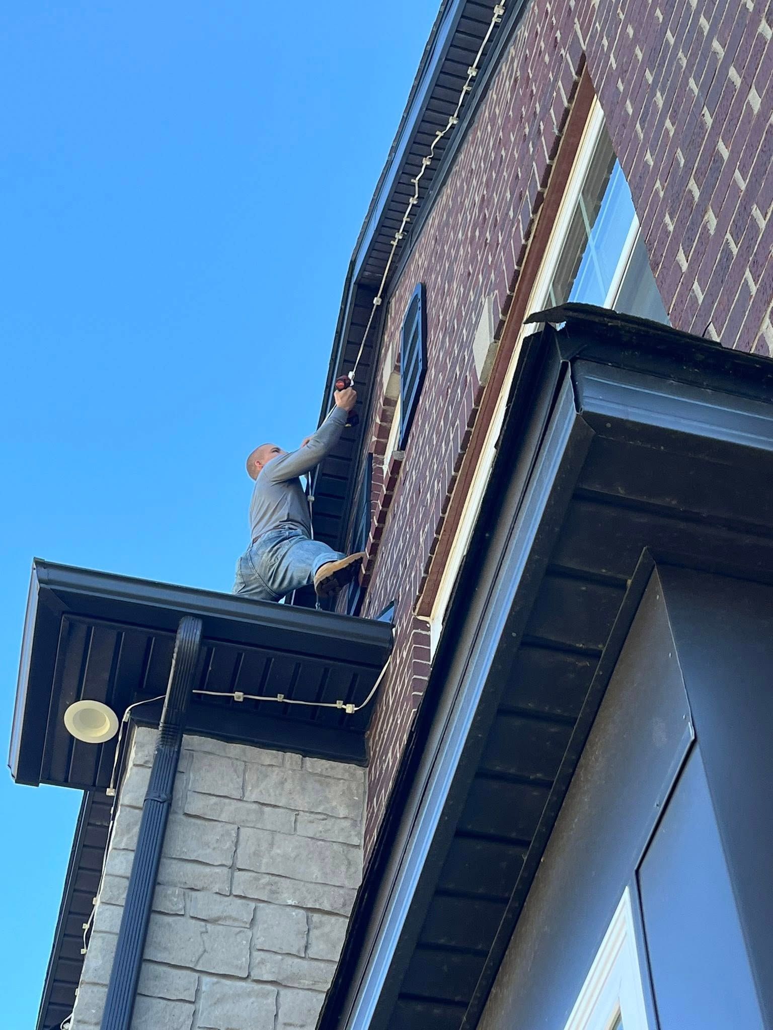 Person on a rooftop attaching lights to a brick building under a clear blue sky.