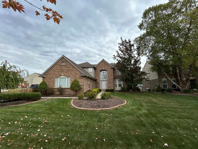 Brick house with a large lawn, circular flower bed, and trees under an overcast sky.