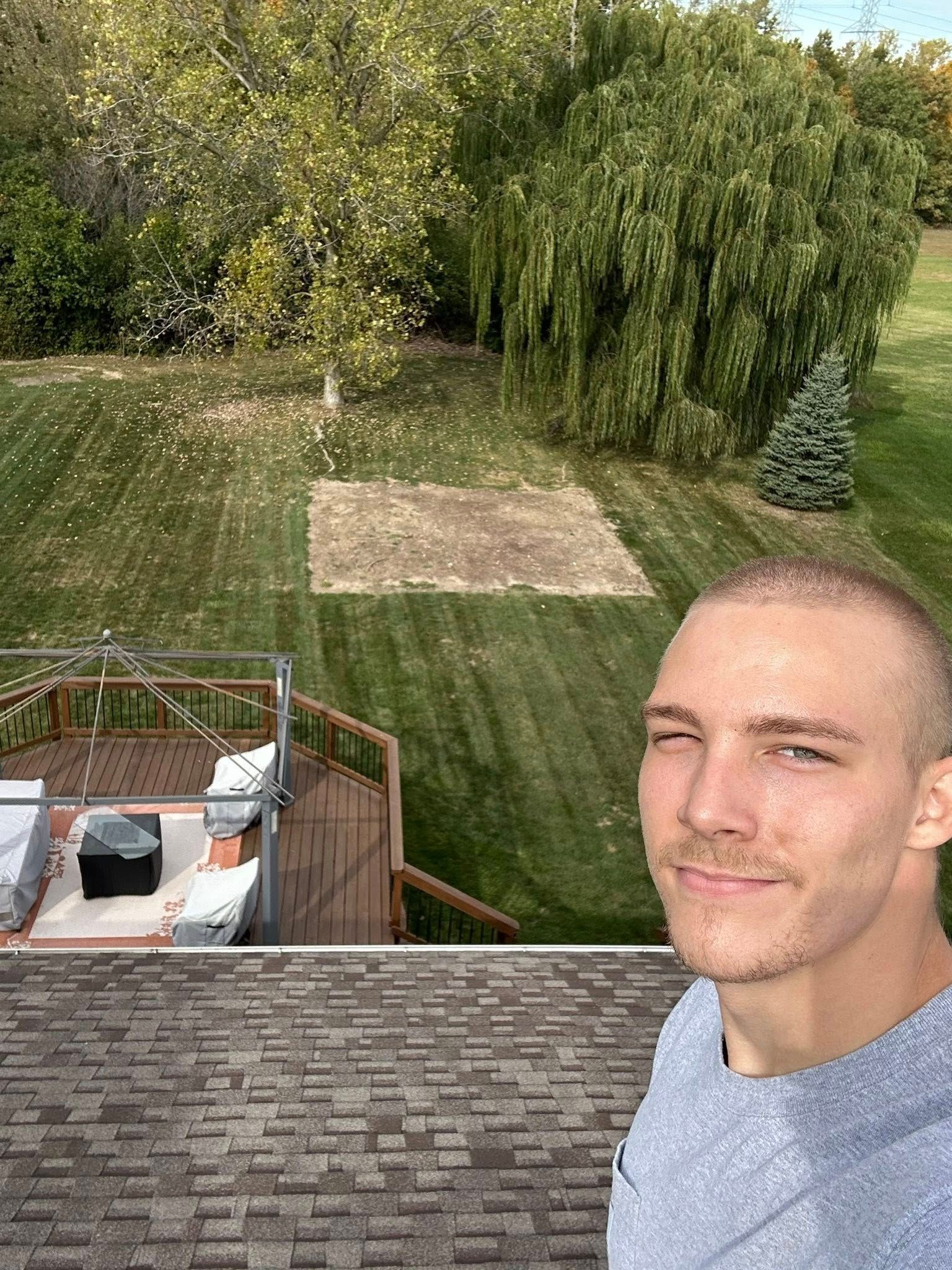 Man taking selfie with backyard view; brown deck, grass, trees, and empty square in yard.