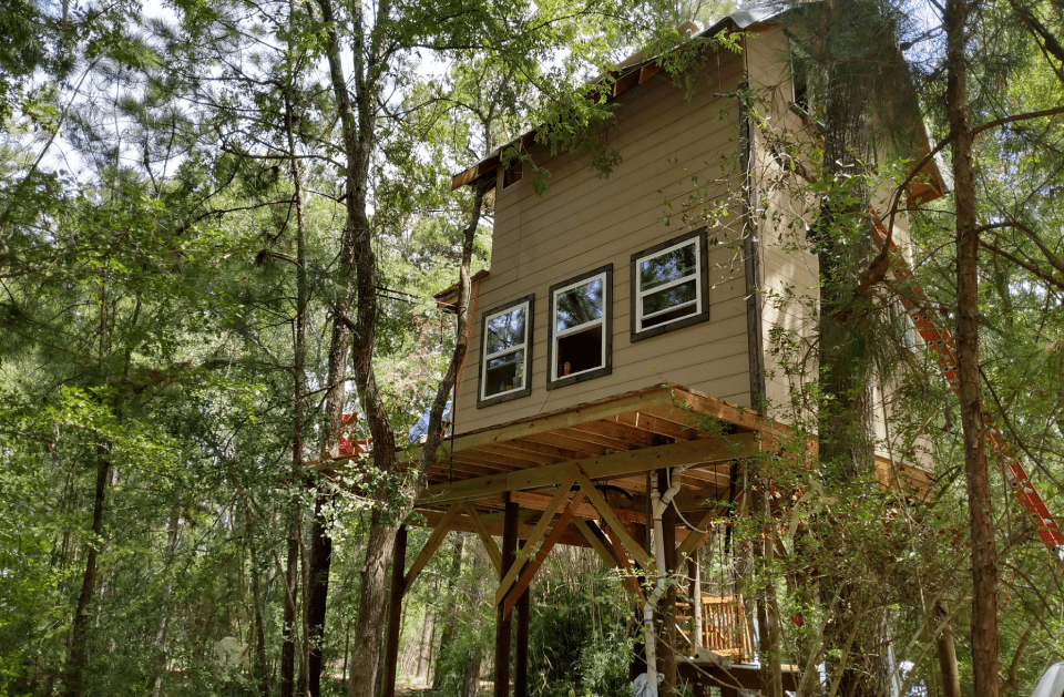 Treehouse with tan siding and wooden supports, nestled in green trees.