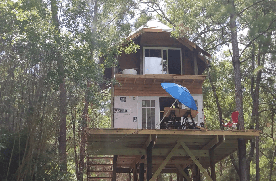 Two-story treehouse with an open deck, blue umbrella, and trees in a sunny outdoor setting.