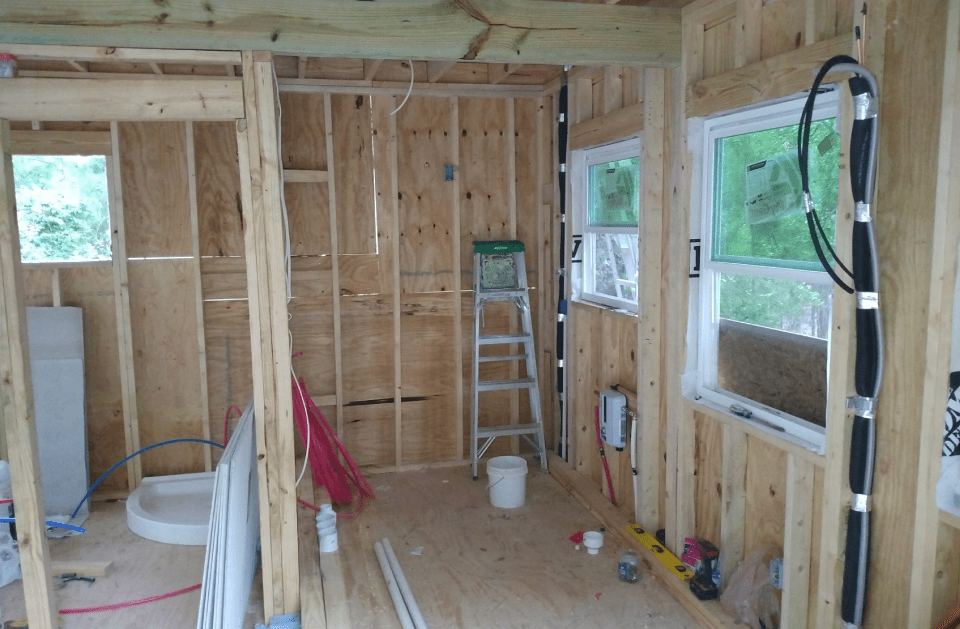 Interior of a small building under construction, showing wooden framing, ladder, and windows.